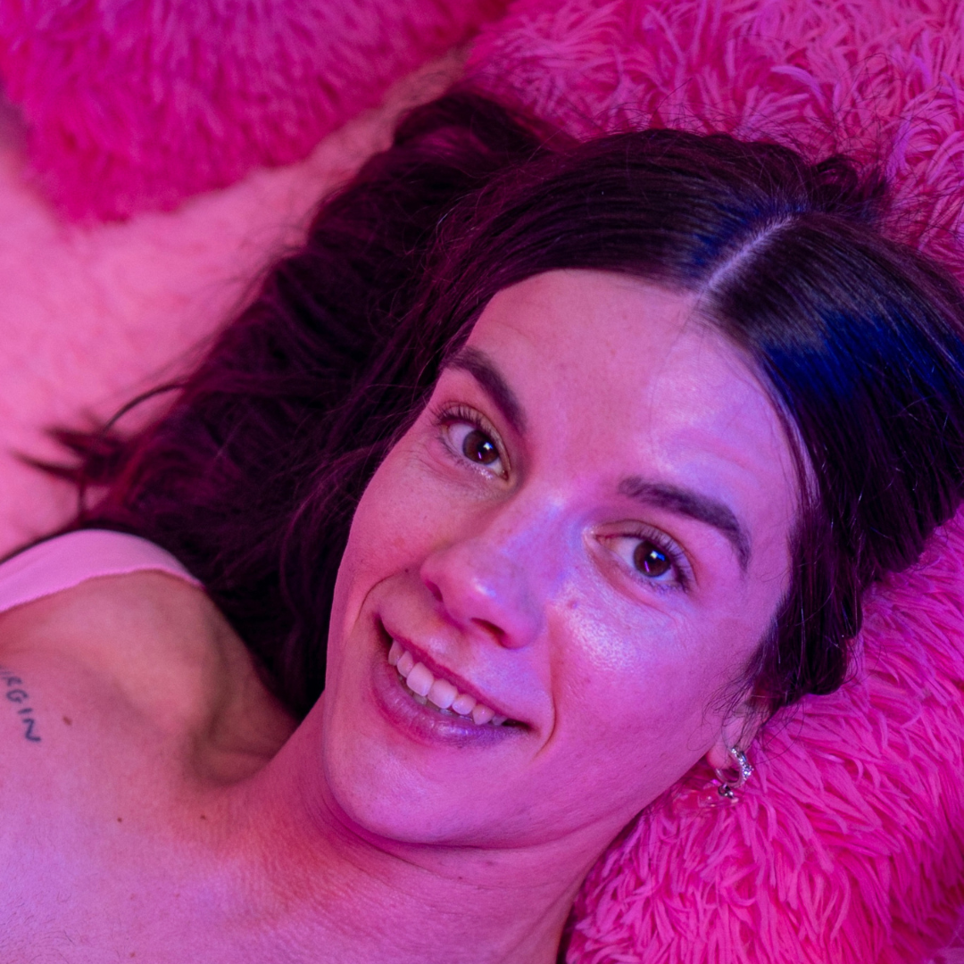 Feminin trans person ligger i flyffy pink puder og smiler til kameraet