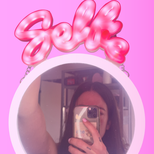 Sissy der tager en selfie inde i et spejl med ringlight hvor der står selfie med neon bogstaver.